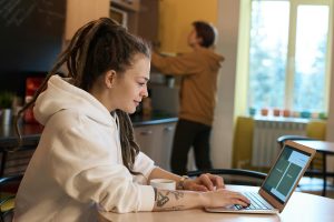 Stock photograph of a freelancer sitting at her kitchen table with her roomate or partner in the background doing something in the kitch. This is to demonstrate the lifestyle of someone trying to build an author platform